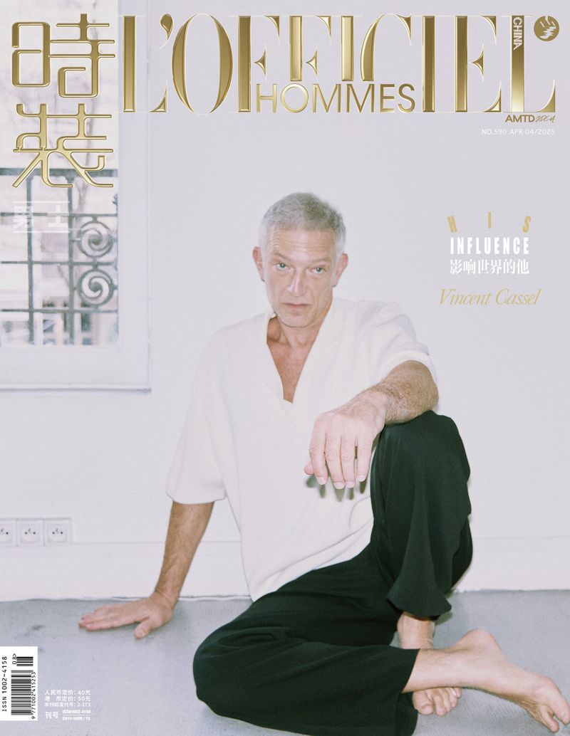 Vincent Cassel poses on the floor in a magazine cover shot. He is wearing a white top and black pants. The background is white.