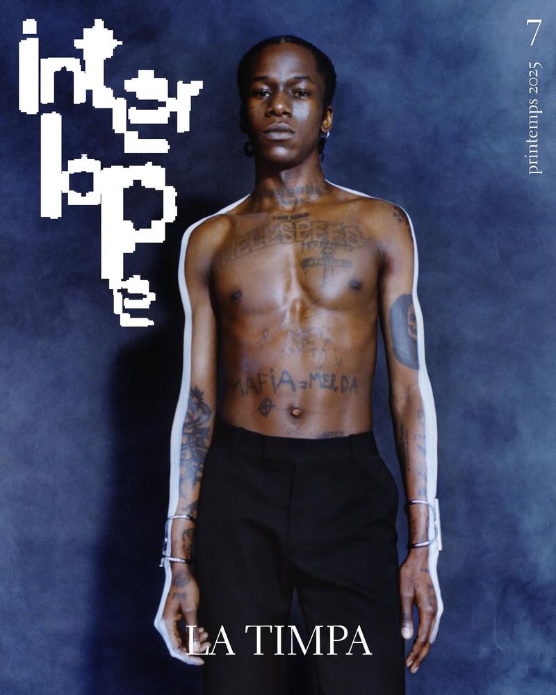 Striking magazine cover featuring a bare-chested Black man with tattoos, jewelry, and black pants against a dark blue gradient backdrop.