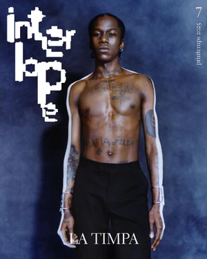 Shirtless young Black man with tattoos wearing dark trousers against a textured dark background for Interlope Magazine