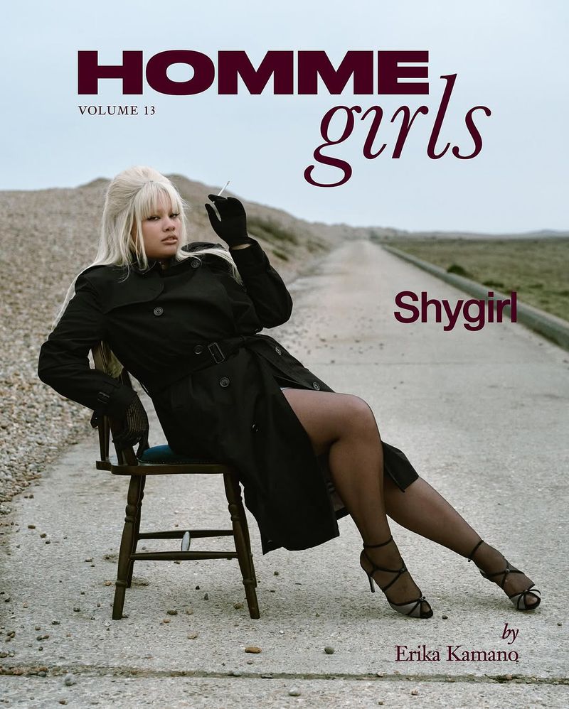 Blonde woman in black trench coat poses seated in chair on outdoor road holding cigarette for magazine cover shoot.