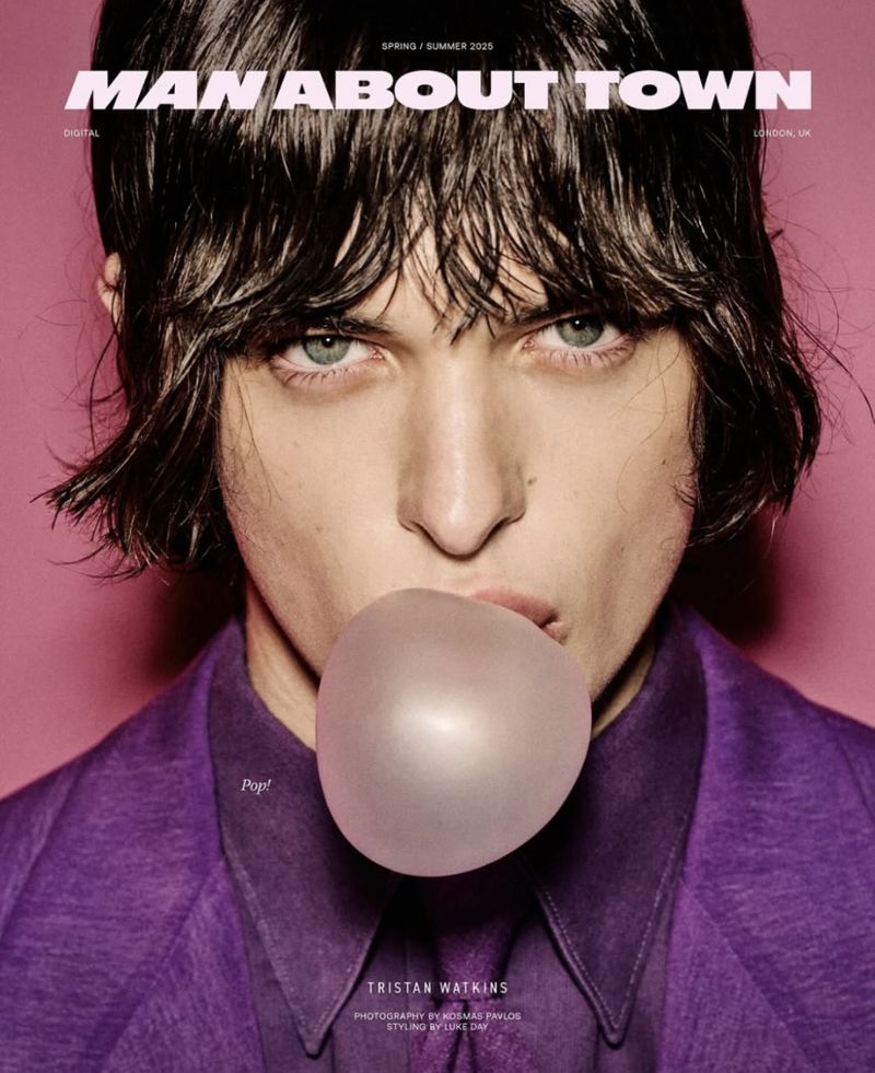 Magazine cover shot of a young adult male model blowing a bubble with bubblegum, wearing a purple suit against a pink background.