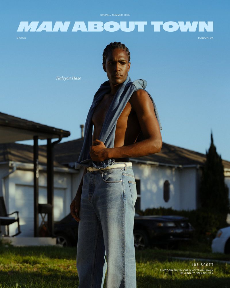 Portrait of a shirtless Black male model wearing jeans and blue jacket outdoors in front of a house on a sunny day.