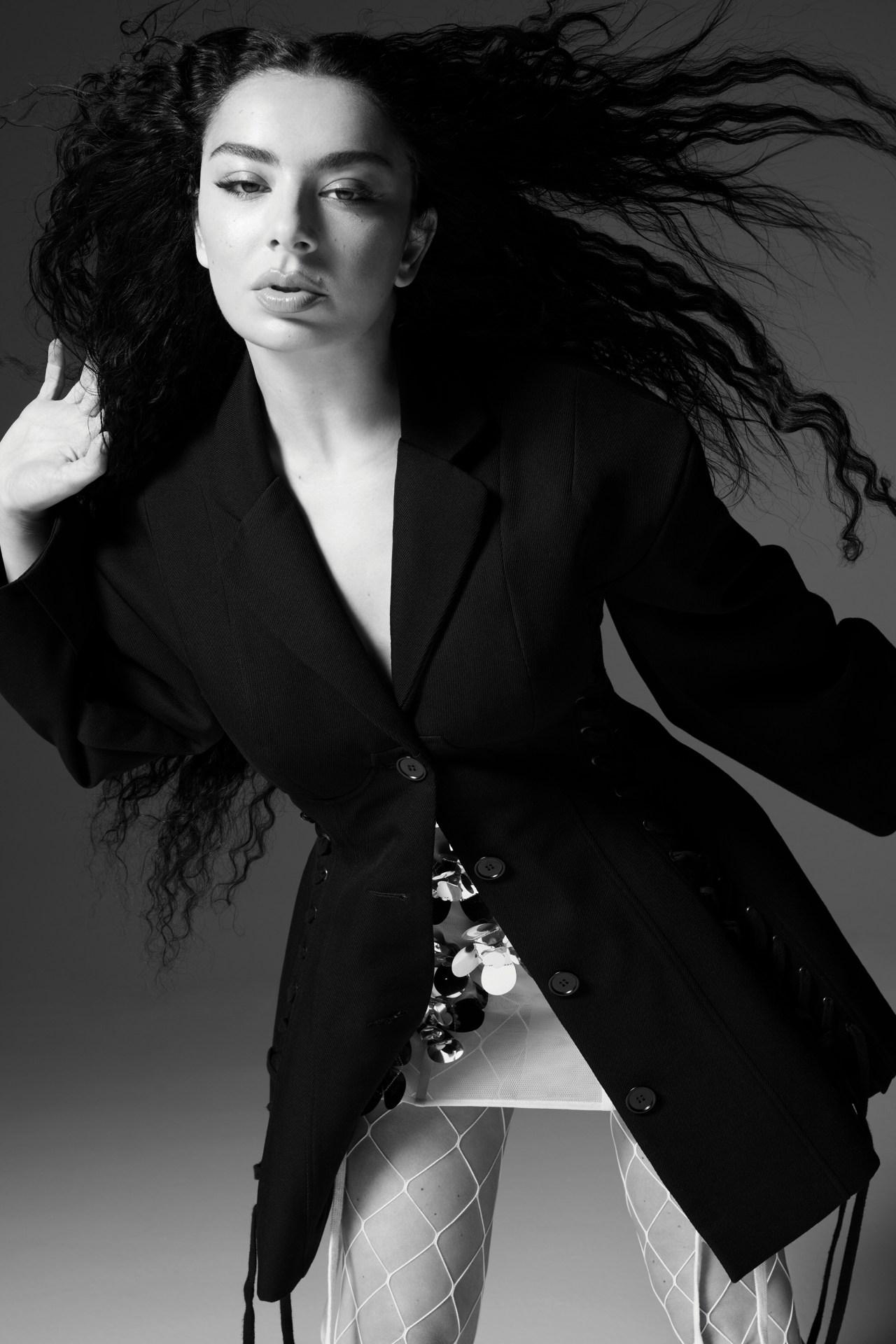 Charli XCX in a black blazer and fishnet tights with long dark hair blowing in a studio portrait for Vogue Australia.