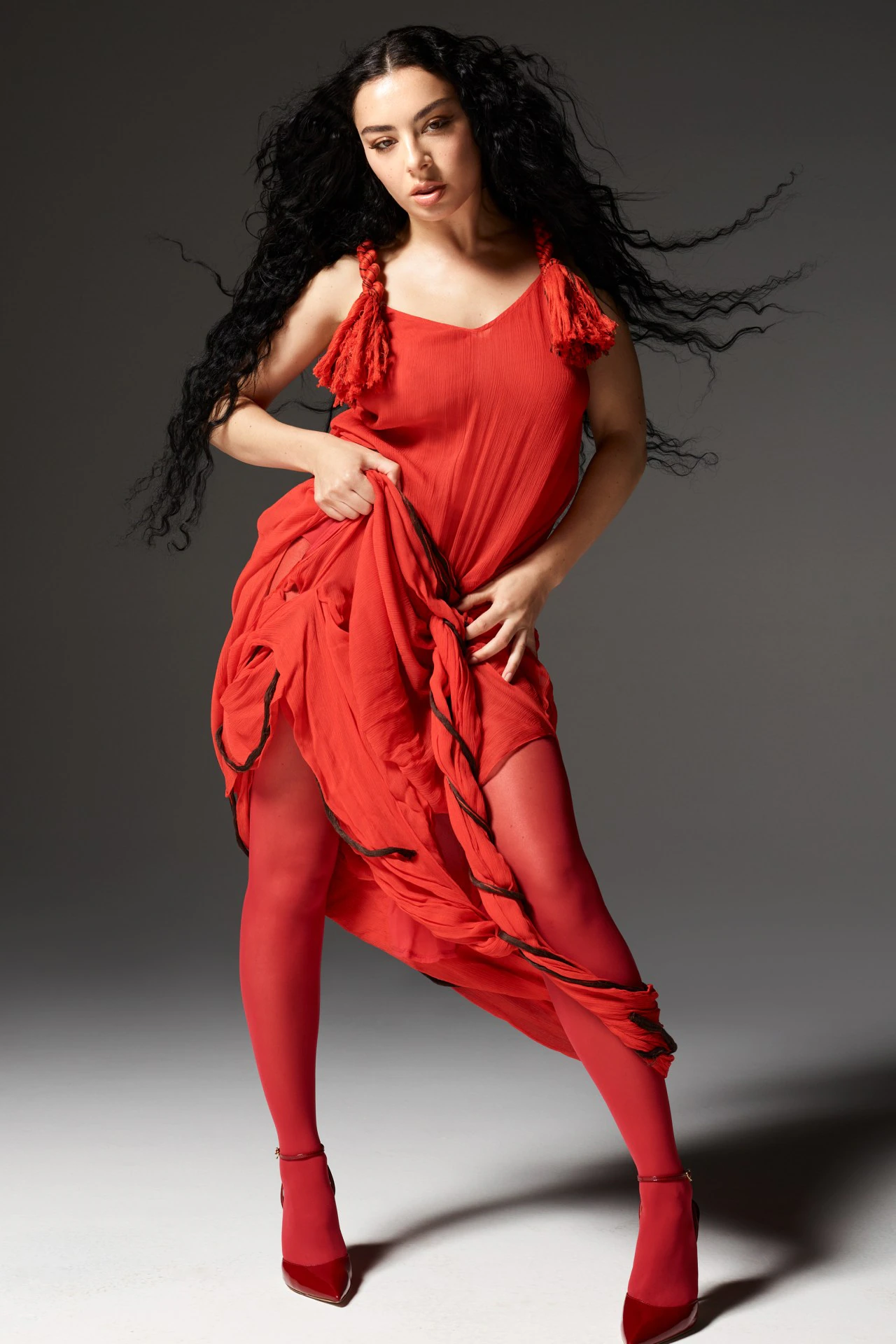 Charli XCX in a red dress and matching tights with flowing dark hair against a grey background