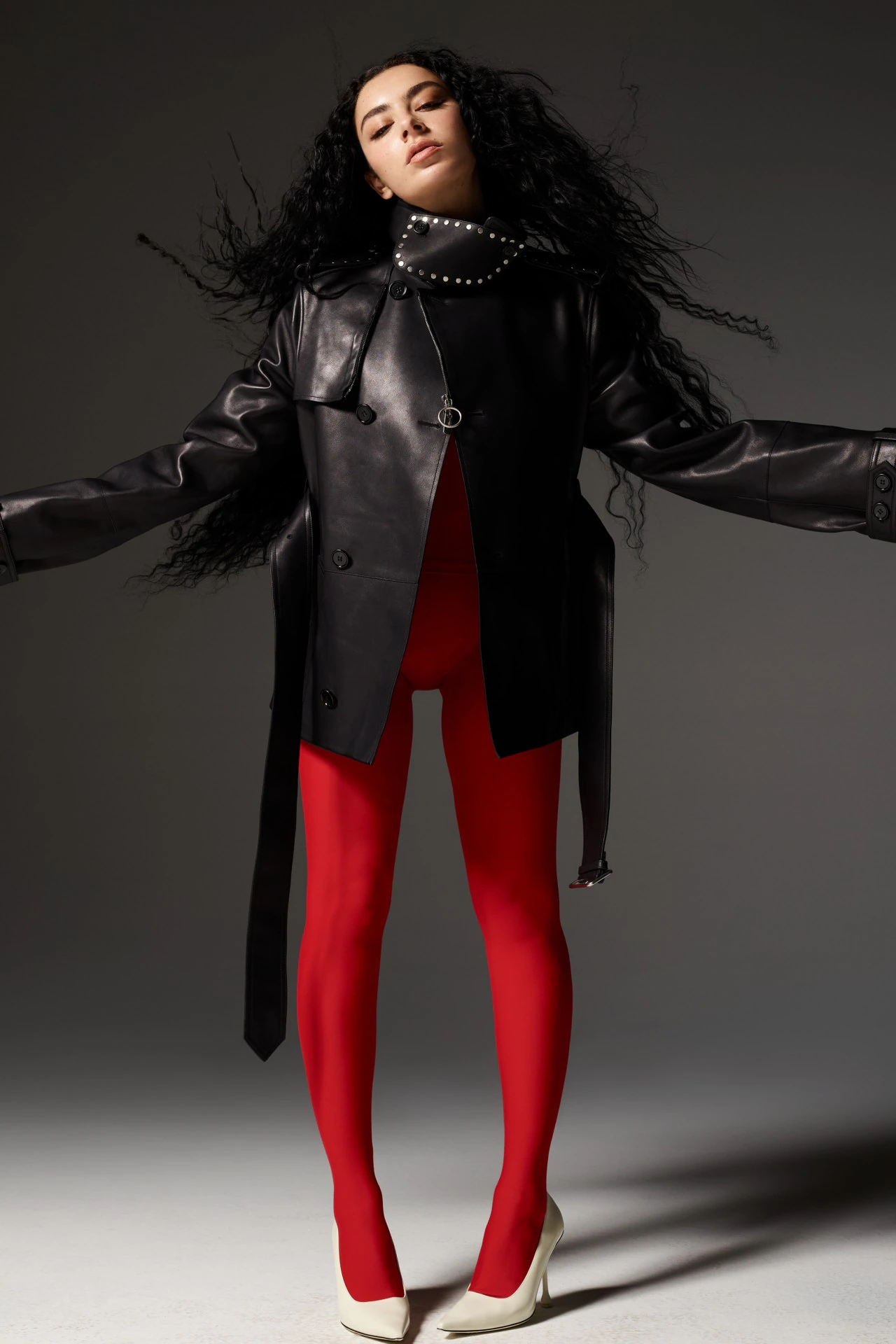 Charli XCX in a black leather jacket and red leggings against a grey background