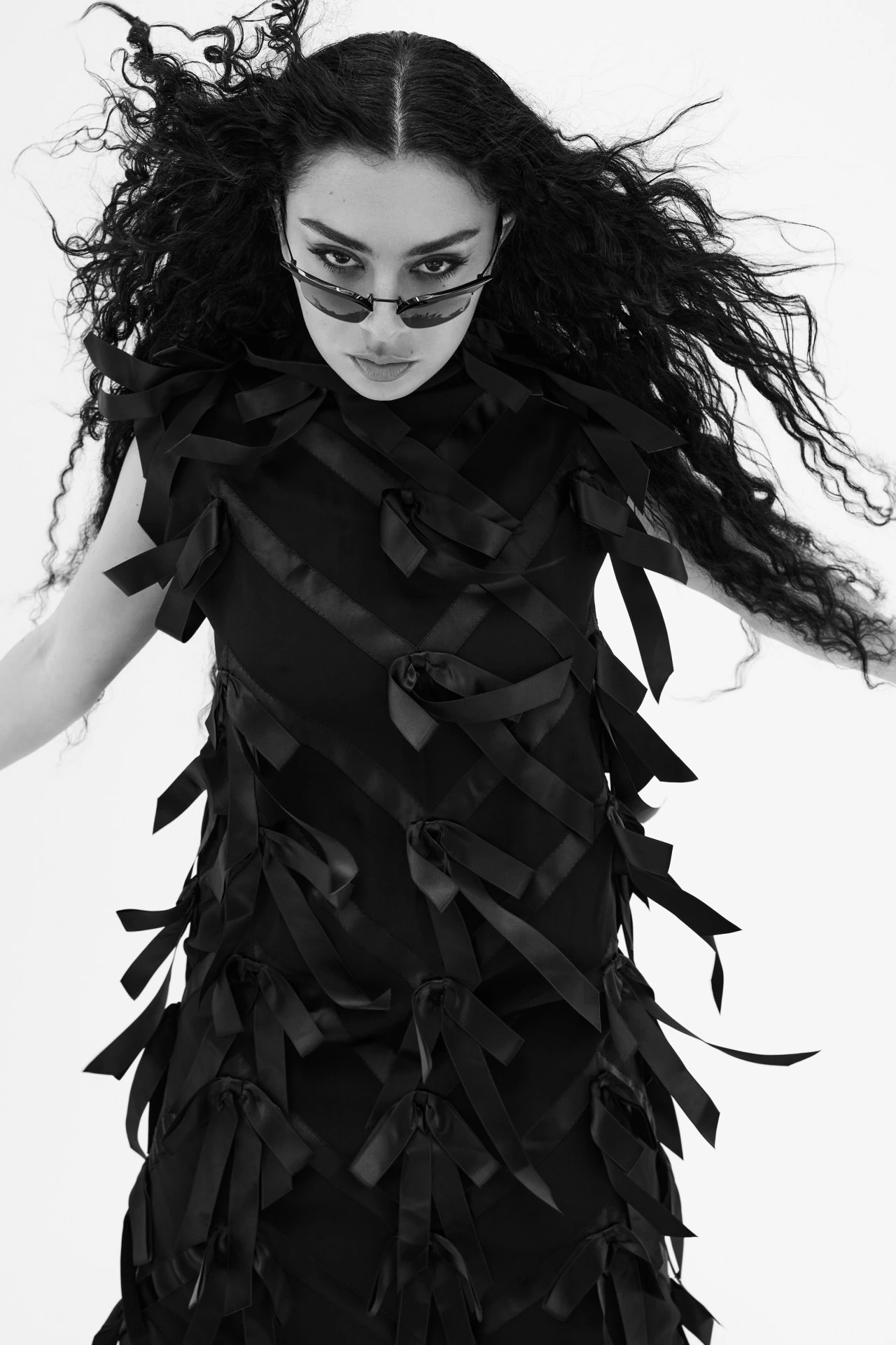 Charli XCX wearing a black ribbon-adorned dress and sunglasses against a white background