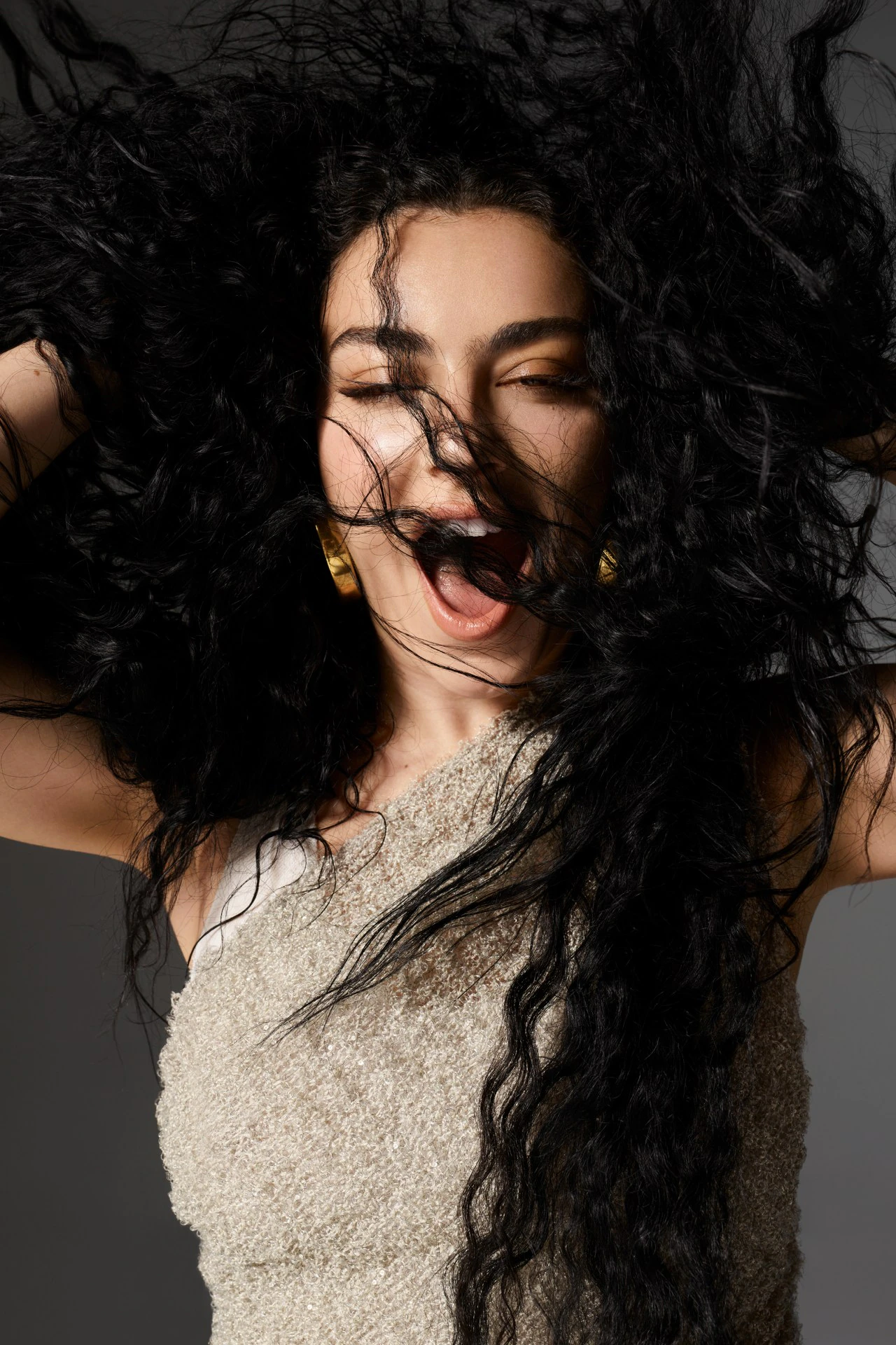 A portrait captures a woman with dark, voluminous hair, eyes closed, mouth open, wearing a textured top against a solid gray backdrop.