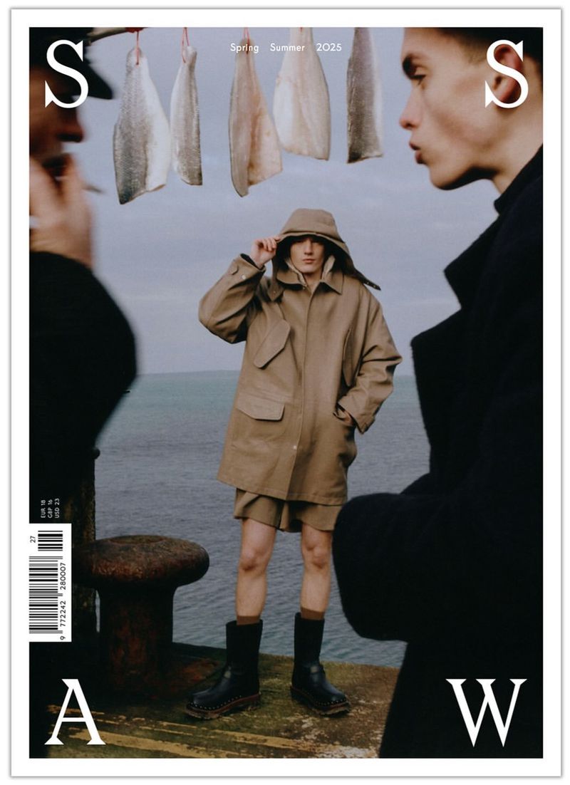 Magazine cover featuring a model in a hooded beige jacket between two other models and suspended fish against a seaside backdrop.