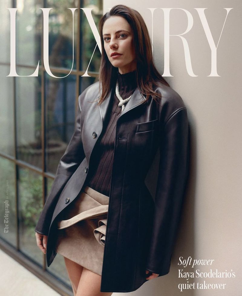 Kaya Scodelario poses in a stylish leather blazer, black turtleneck, and tan skirt against a neutral wall with a glass window.