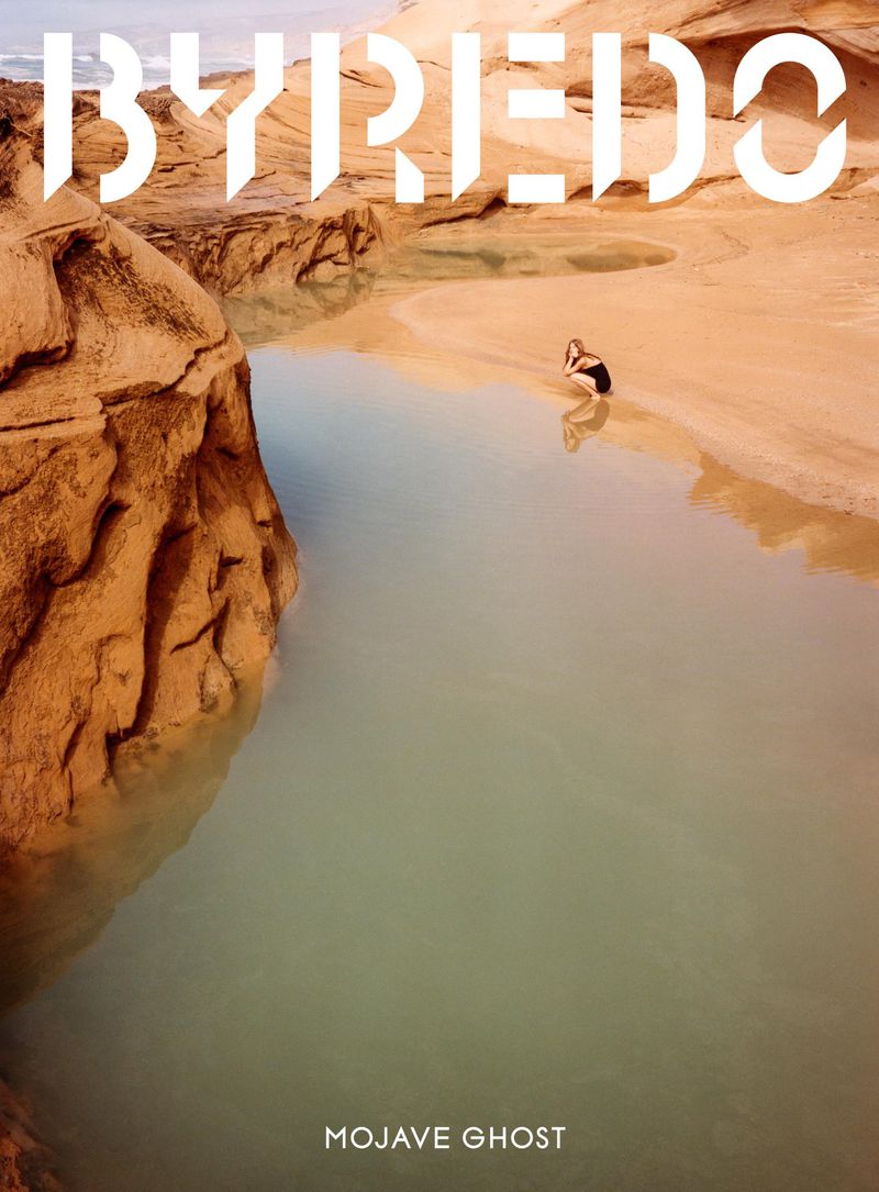 A woman in a black swimsuit stands in shallow water between tan rock formations, under a logo for BYREDO and the text "MOJAVE GHOST."