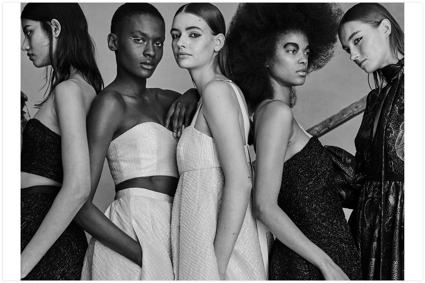 Black and white studio shot featuring five female fashion models in elegant dresses, standing close together in a neutral setting.