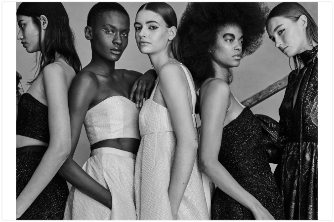 Black and white studio shot featuring five female fashion models in elegant dresses, standing close together in a neutral setting.