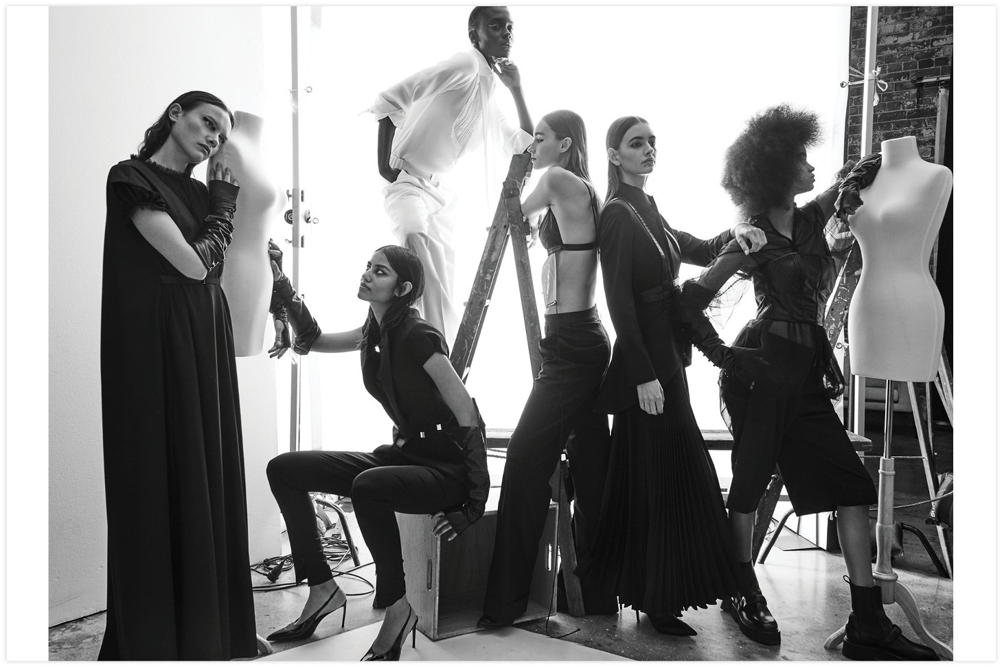 Black and white studio shot of multiple models in dark clothing posing around mannequins and a ladder, fashion editorial.
