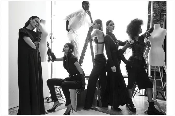 Black and white studio shot of multiple models in dark clothing posing around mannequins and a ladder, fashion editorial.