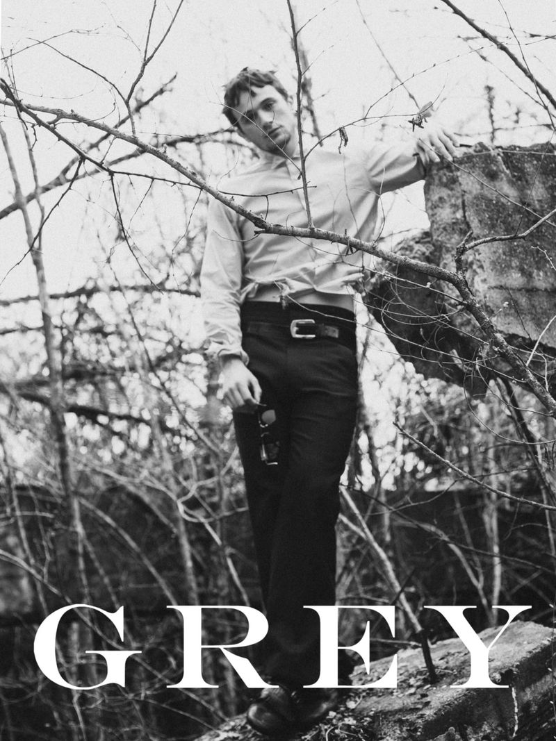 Monochrome fashion shot: a male model in a shirt and trousers poses among bare branches and concrete rubble, creating an editorial aesthetic.
