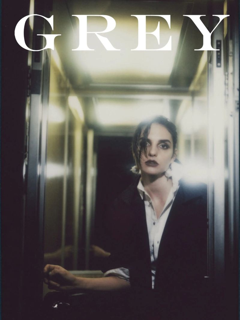 Portrait of a woman in a black blazer and white shirt stands in a hallway, illuminated by soft, diffused light, with the word 'GREY' above.