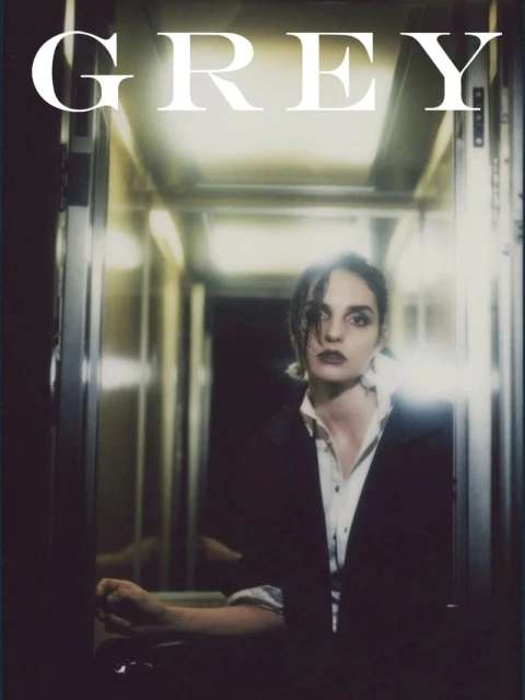 Portrait of a woman in a black blazer and white shirt stands in a hallway, illuminated by soft, diffused light, with the word 'GREY' above.