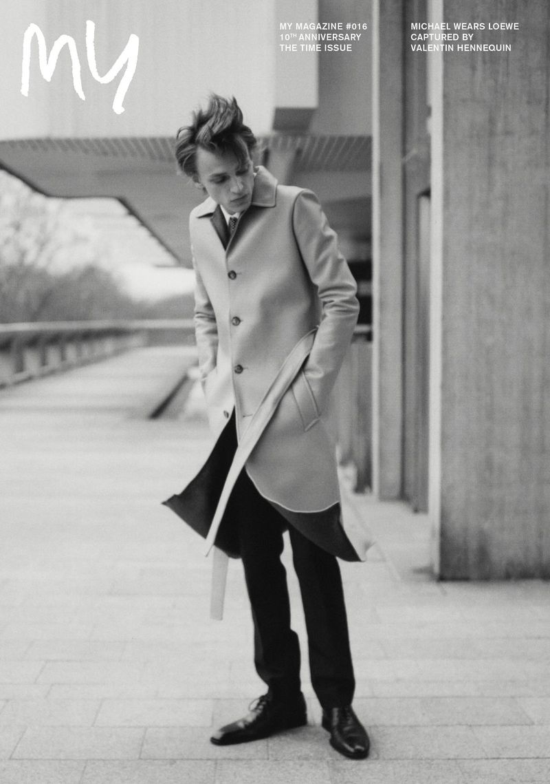 A monochromatic fashion editorial presents a slender man in a trench coat, captured outdoors in a full shot with the magazine logo.