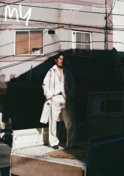 Man standing in the back of a white truck wearing an off-white coat and matching trousers against a building exterior.