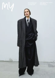 Model wearing oversized black patterned coat and suit against a plain white studio wall