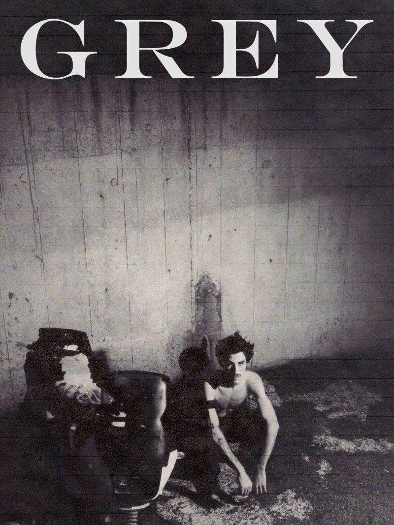 Monochrome portrait of a shirtless man squatting next to a vintage barber chair in a gritty, textured room under bright lighting.