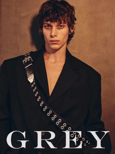 Portrait of a young adult male with brown hair wearing a black blazer and a black strap bag with silver hardware, against a neutral backdrop.