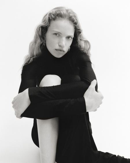 Monochrome portrait of a woman wearing a black turtleneck top, seated with knees drawn up against a seamless white background.