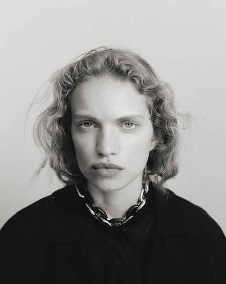 Black and white portrait of a woman with curly blonde hair wearing a black jacket and silver chain link necklace in a studio.