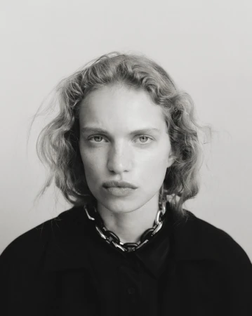 Black and white portrait of a woman with curly blonde hair wearing a black jacket and silver chain link necklace in a studio.
