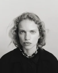 Close-up portrait of a woman with curly hair wearing a dark shirt and a chunky chain necklace against a light background.