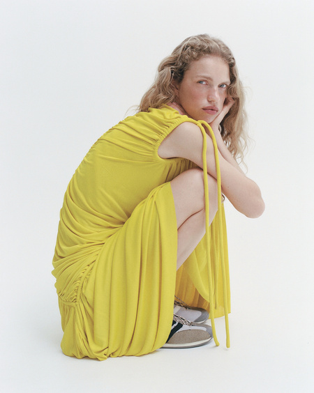 Woman with blonde curly hair crouching in a bright studio wearing a gathered yellow dress and sneakers. Minimalist fashion photography.