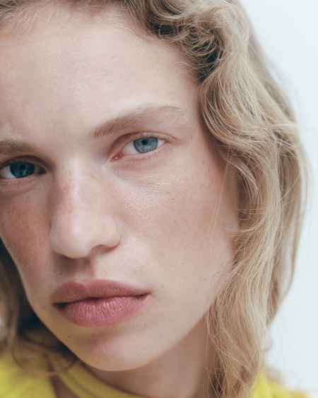 Close-up portrait of a woman with fair skin and blue eyes, framed by curly blonde hair, against a bright neutral background. Beauty and fashion.
