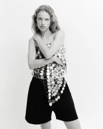 Model with long wavy hair wearing a metallic sequined strapless top and dark shorts against a bright white background.
