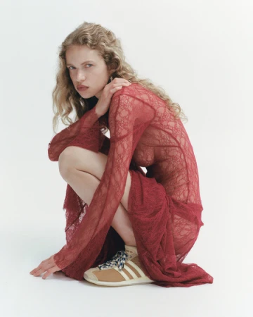 Woman with blonde curly hair poses in a red sheer dress and sneakers against a white backdrop in a studio with soft lighting.