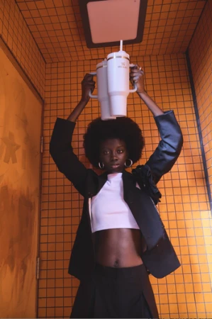 Woman in blazer holds industrial-style cups overhead in an orange tiled room with geometric fixtures and black wire framing.