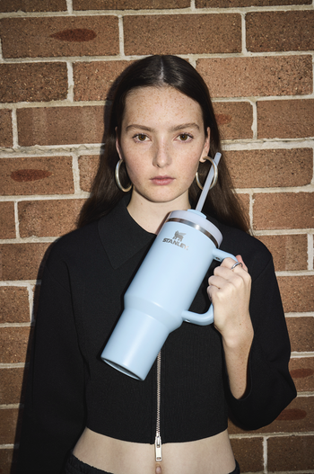 Young woman with freckles poses with a light blue Stanley cup and straw in front of a textured brick wall in a medium shot.