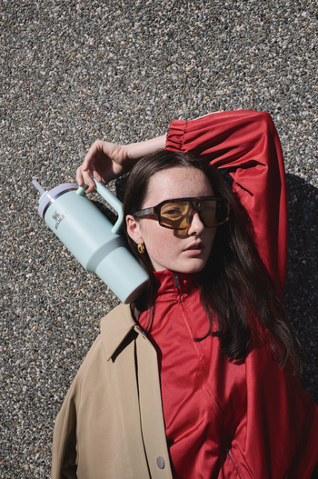 Young woman with sunglasses poses outdoors with a large pale blue water bottle, wearing a color-blocked beige and red jacket against a textured wall.