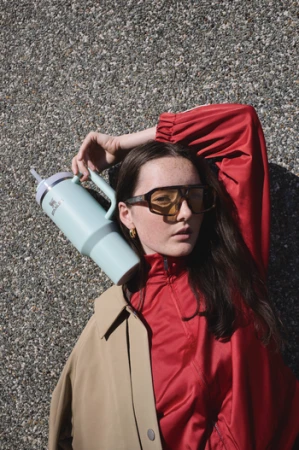 Young woman with sunglasses poses outdoors with a large pale blue water bottle, wearing a color-blocked beige and red jacket against a textured wall.
