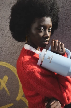 An African American woman holds a light blue Stanley tumbler with a straw in front of a textured wall with graffiti.