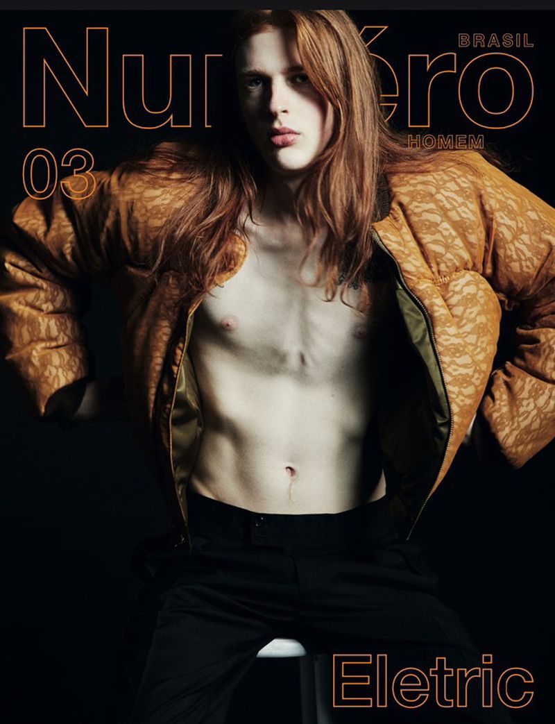Magazine cover featuring a shirtless male model with auburn hair, wearing an open tan jacket and black pants against a dark background.