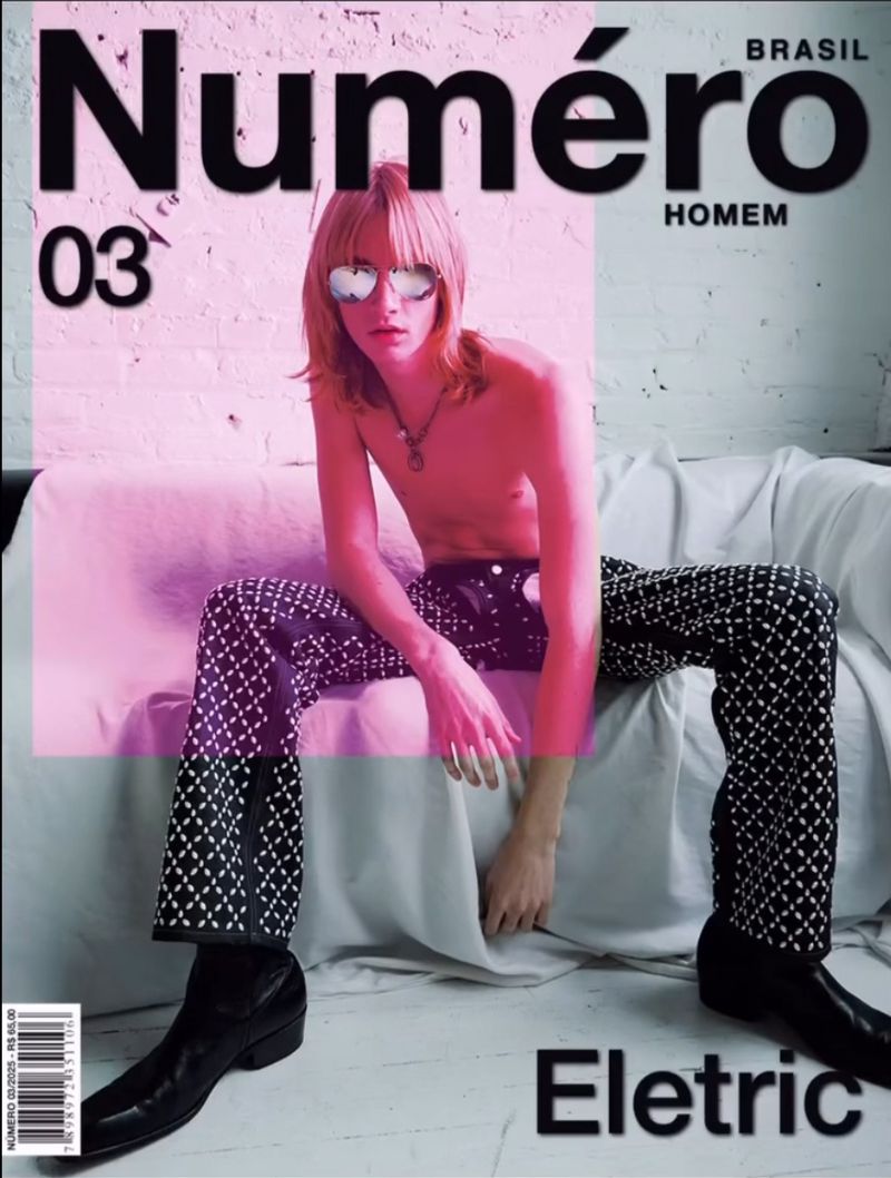 Magazine cover of a shirtless young adult male in decorative black pants and sunglasses, seated on a white couch in front of a white wall.