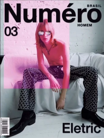 Magazine cover of a shirtless young adult male in decorative black pants and sunglasses, seated on a white couch in front of a white wall.