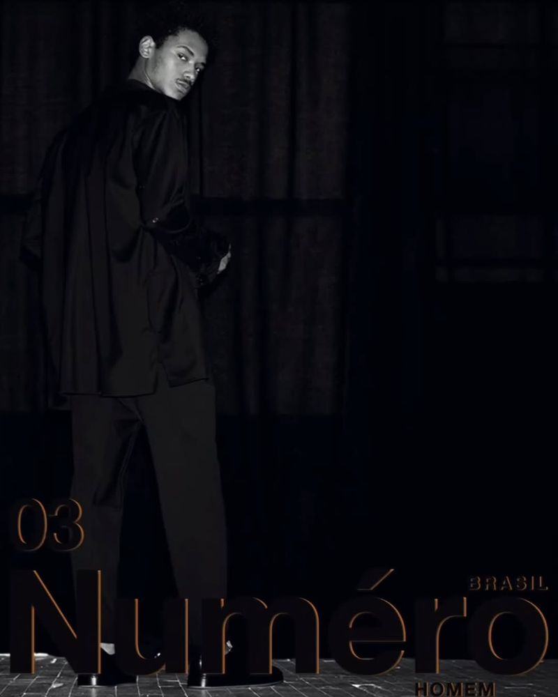An adult male model poses indoors wearing black satin shirt and pants, looking over his shoulder in low-key, dramatic lighting.