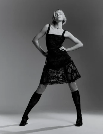 Blonde woman in a black lace dress and knee-high socks poses in a photo studio. Fashion photography in black and white.
