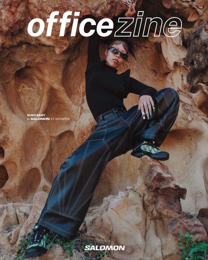 Magazine cover shot of adult female model in black top and wide-leg pants, posed against a rocky background in outdoor lighting.