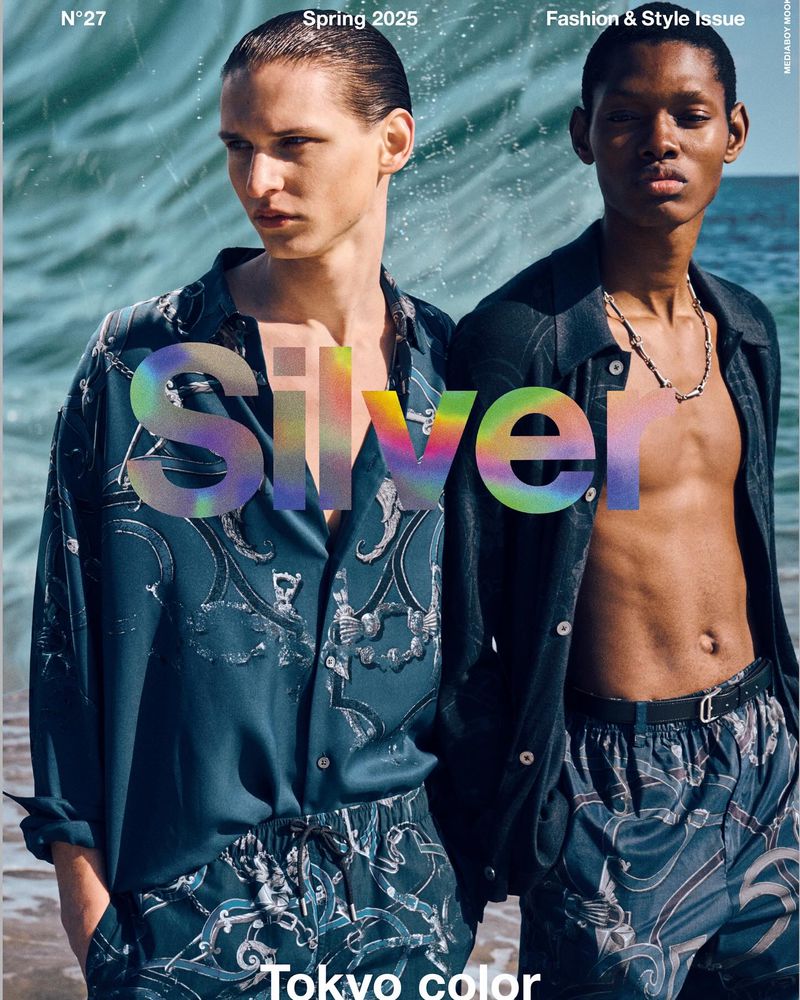 Two models pose against an ocean backdrop in matching patterned shirts and shorts for a fashion magazine cover.