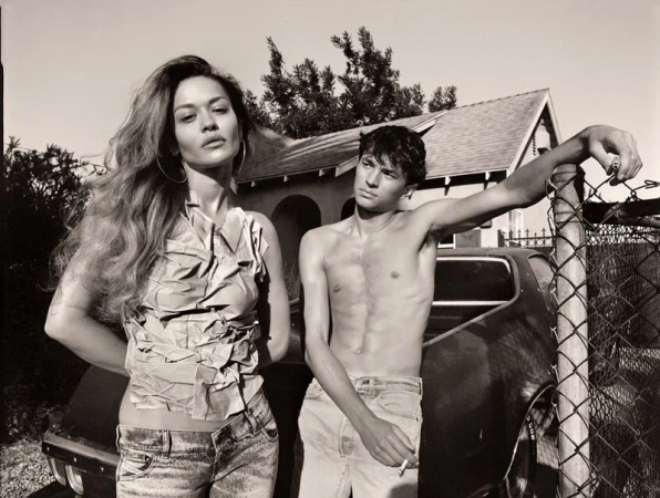 Monochrome portrait of a woman with long hair standing next to a shirtless man leaning on a fence and car, under a bright sky.
