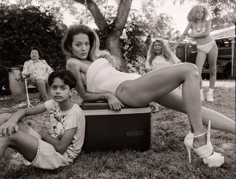 Black and white outdoor shot of a woman in a bodysuit and high heels, sitting on a cooler, along with a child and three older women.