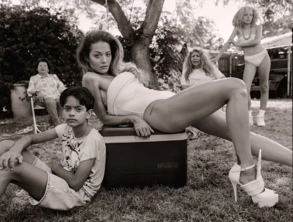 Black and white outdoor shot of a woman in a bodysuit and high heels, sitting on a cooler, along with a child and three older women.