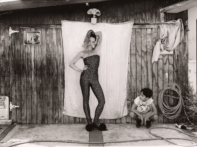 Black-and-white image of a woman in a patterned jumpsuit and a child squatting in front of a wooden wall, garden hose, and a white sheet.